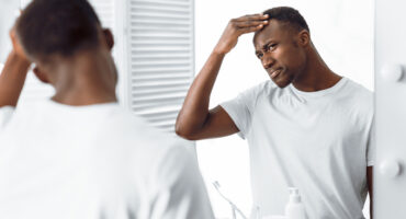 Black man in mirror examining his own hair line in a white t shirt.