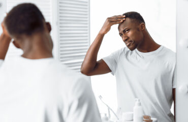 Black man in mirror examining his own hair line in a white t shirt.