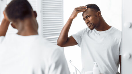 Black man in mirror examining his own hair line in a white t shirt.